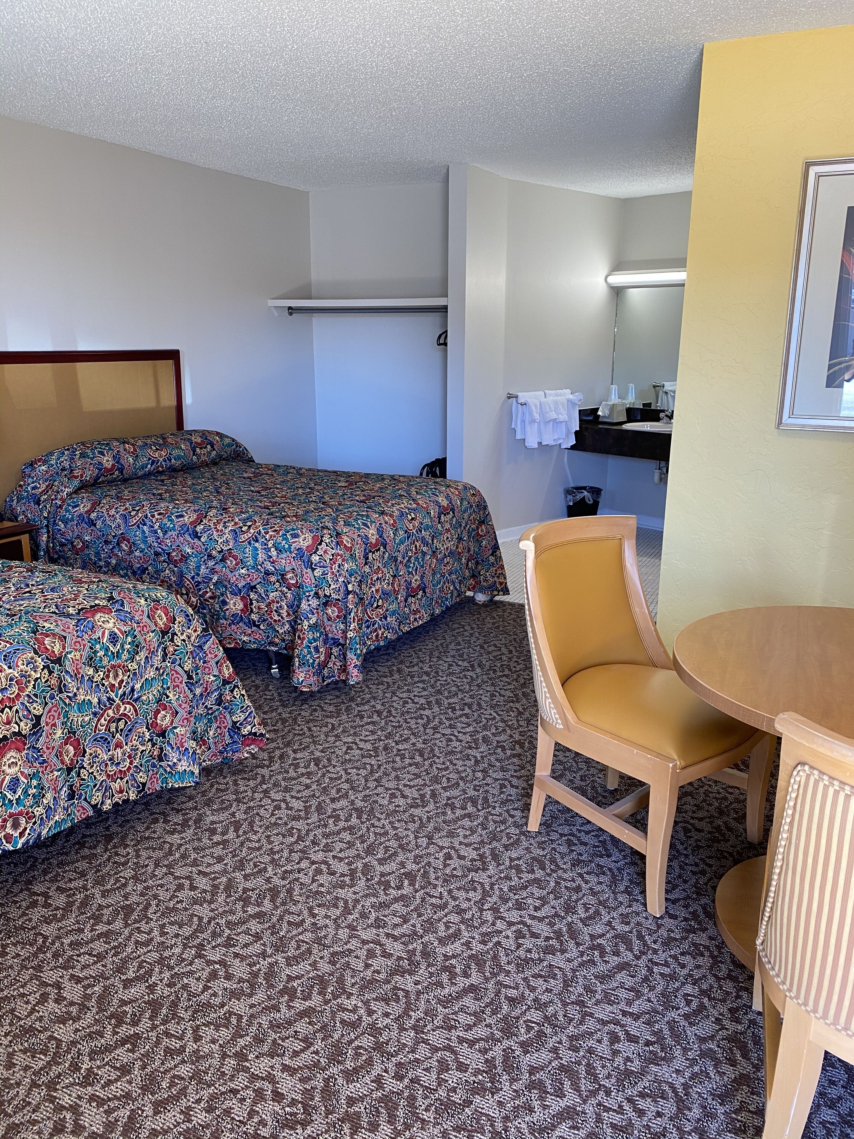 A hotel room with two beds, a chair, a small table, a bathroom with a sink and mirror, and a carpeted floor.