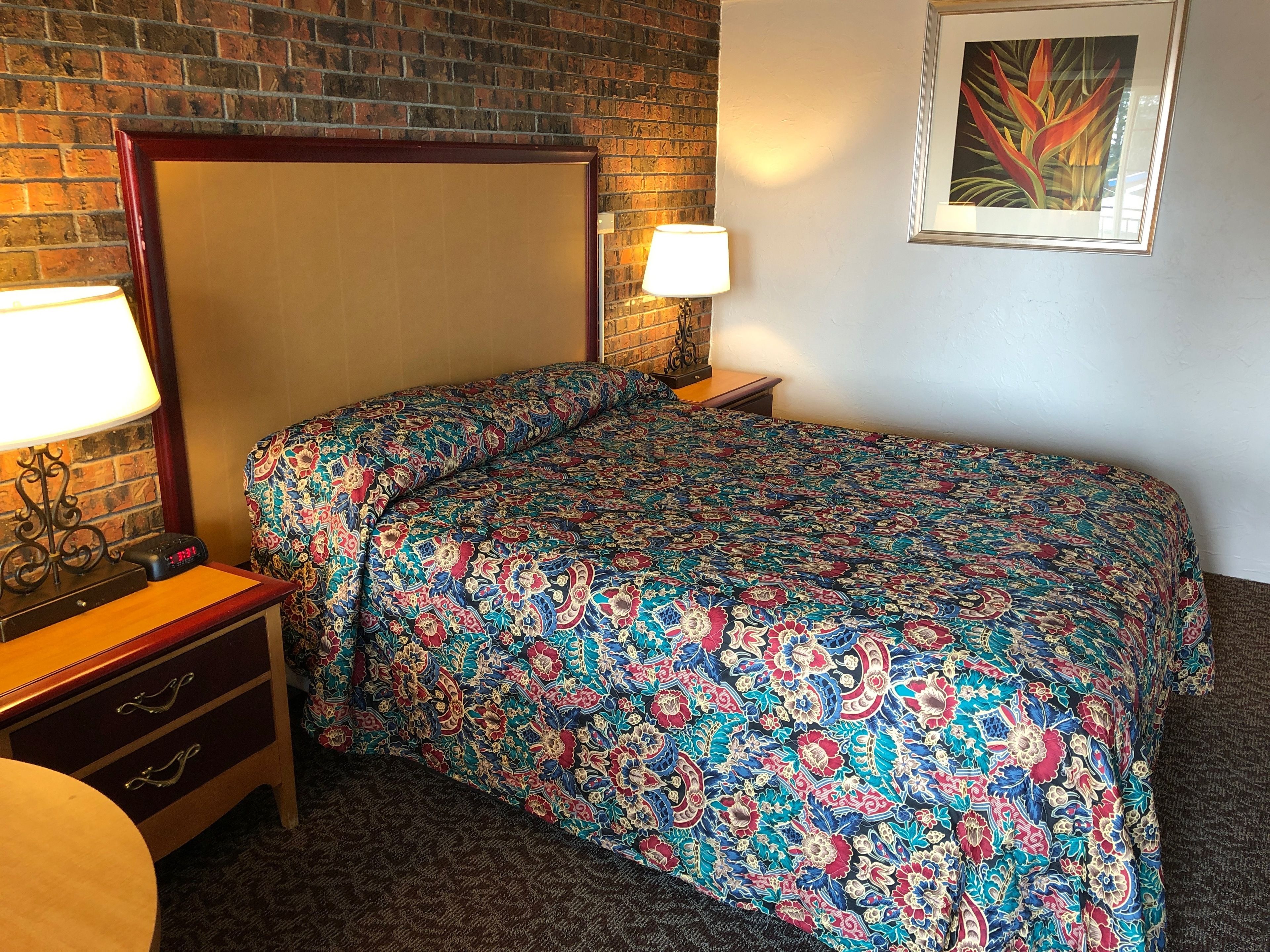 A hotel room with a brick wall, a bed with a colorful floral bedspread, a nightstand with a lamp, and a small table with a clock.