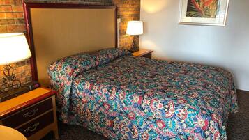A hotel room with a brick wall, a bed with a colorful floral bedspread, a nightstand with a lamp, and a small table with a clock.