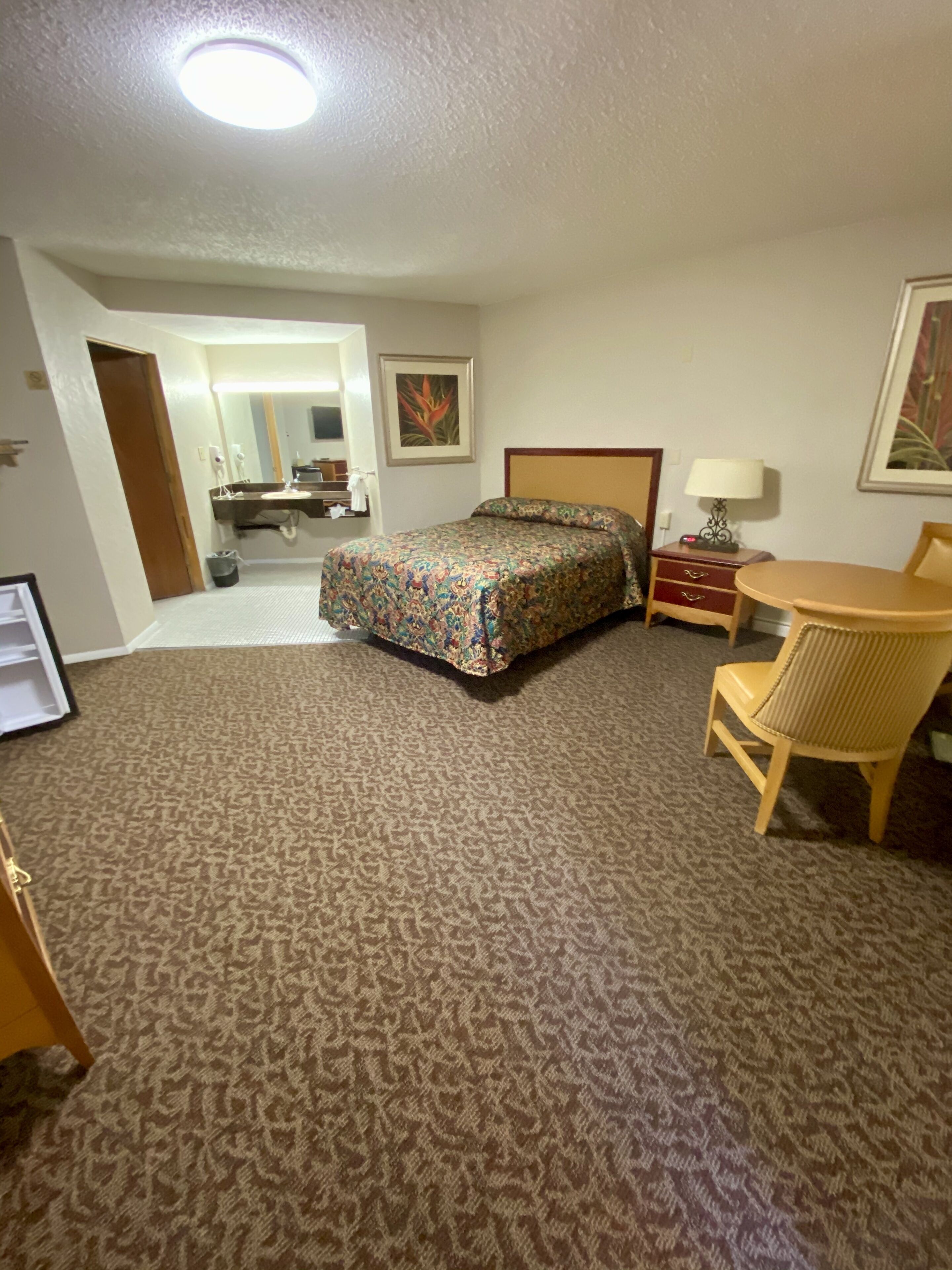 A hotel room with a bed, a desk, a chair, a lamp, and a bathroom visible through an open door.