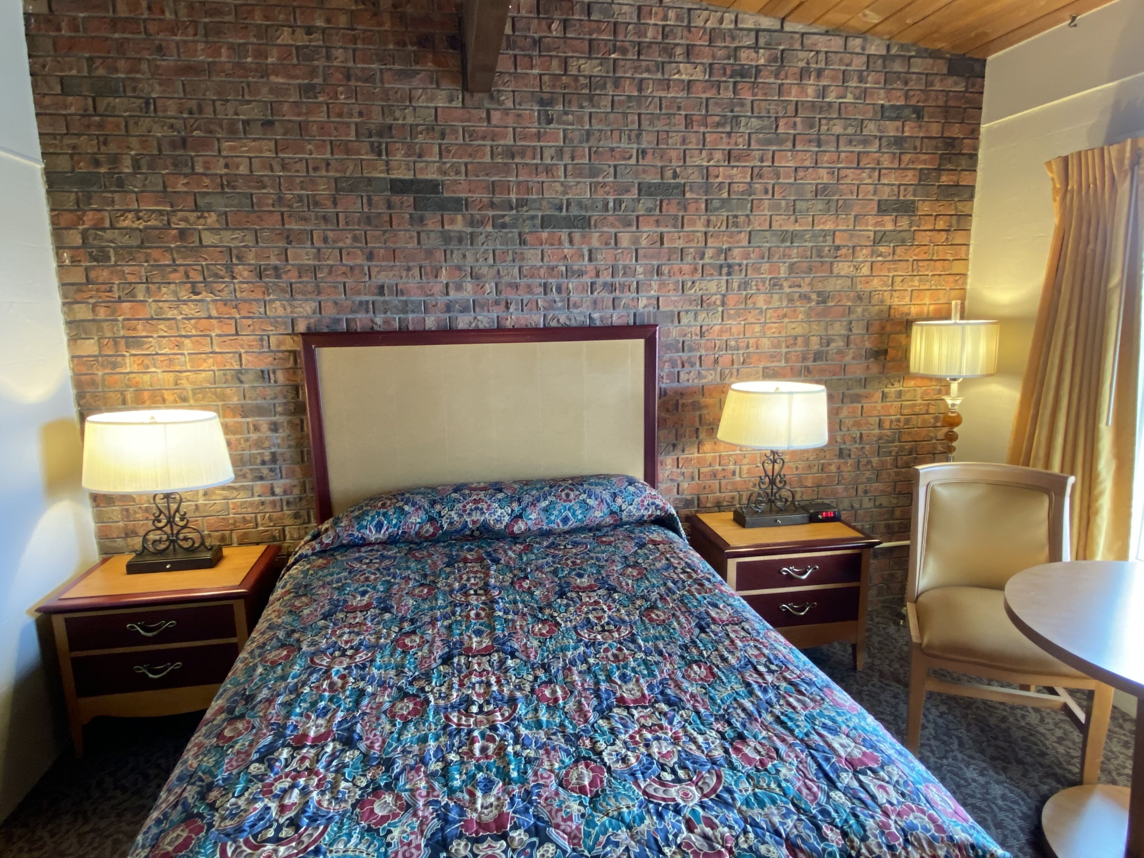 Ein Hotelzimmer mit Backsteinwand, einem Bett mit gemustertem Bettzeug, zwei Nachttischen mit Lampen, einem Sessel und einem kleinen Tisch.