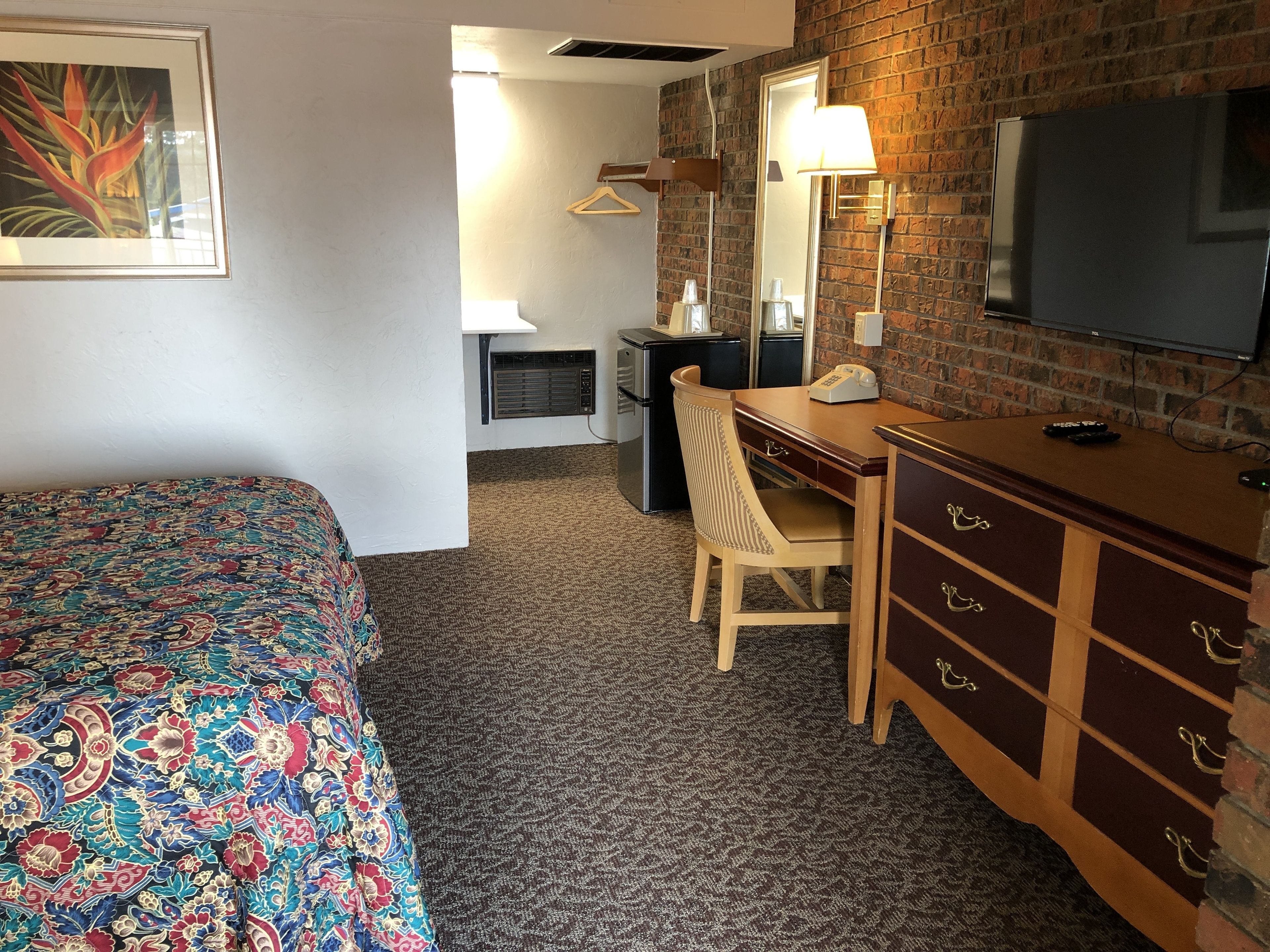 Ett hotellrum med en säng, ett skrivbord, en byrå, en spegel, en tv och ett fönster med persienner.