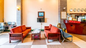 Un vestíbulo con sofás rojos, una mesa de centro, un televisor y una recepción con relojes y un menú.
