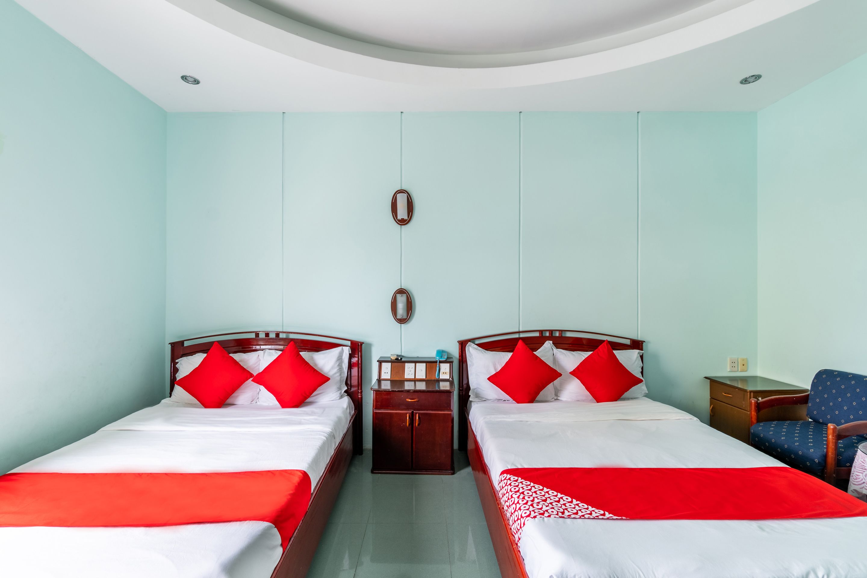 A hotel room with two beds, each with red and white bedding, a small wooden nightstand, a desk with a chair, and a light blue wall.