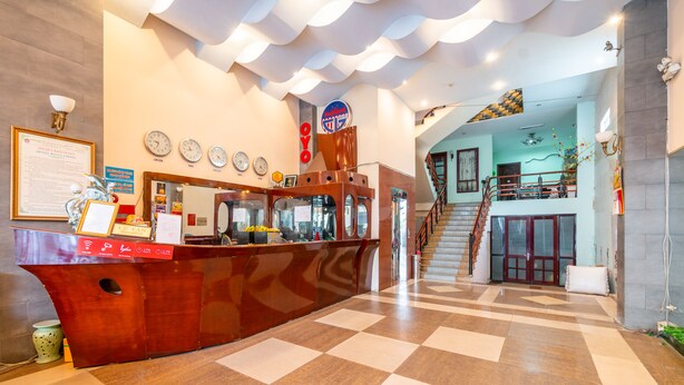 Un vestíbulo de hotel con mostrador de recepción, relojes y una escalera que sube a un piso superior.