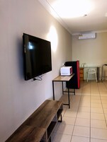 Habitación con televisor de pantalla plana montado en la pared, un banco de madera, una mesita con microondas y una zona de kitchenette con refrigerador y una escalera.