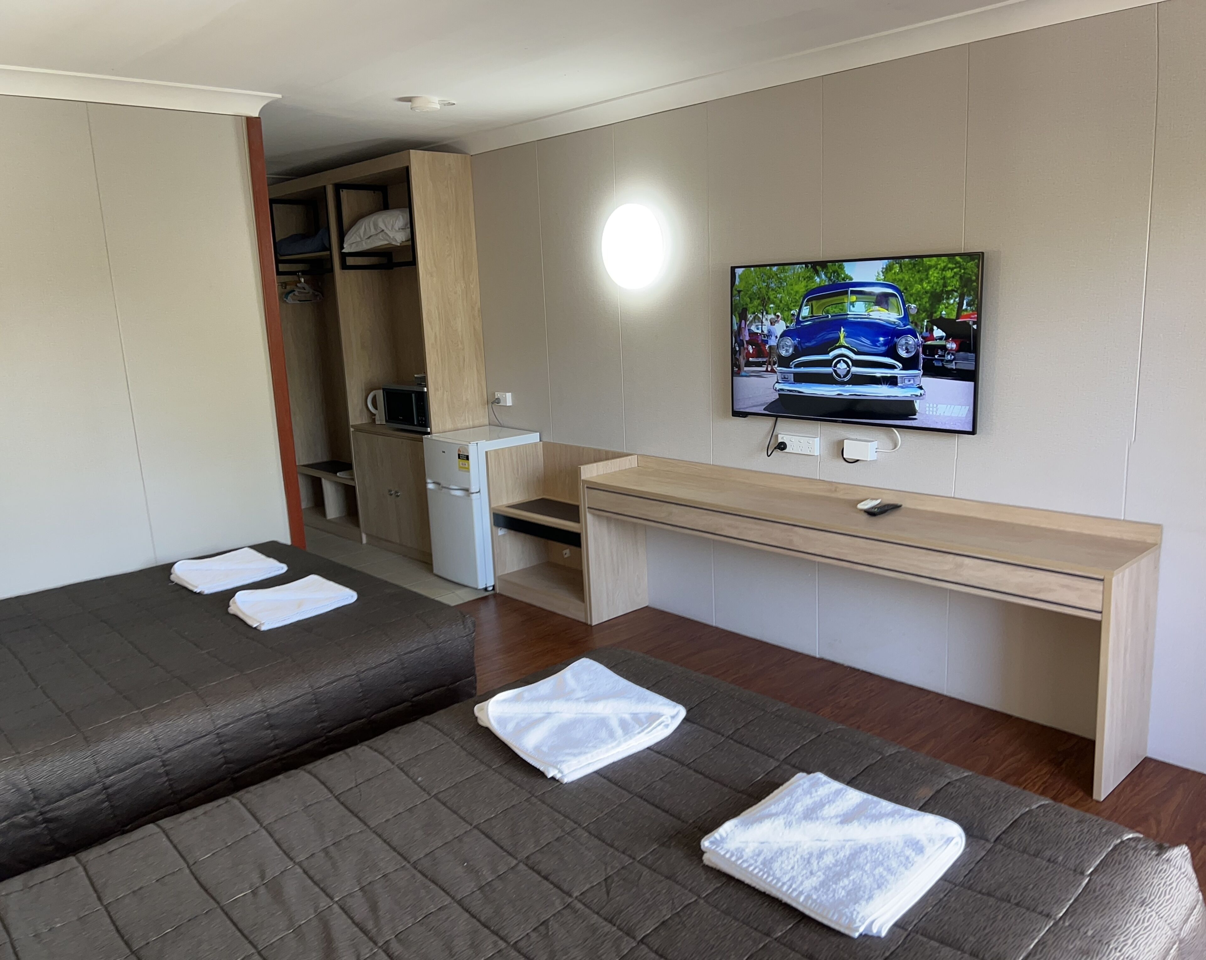 A compact hotel room with two beds, a small kitchenette, a TV, and a desk.