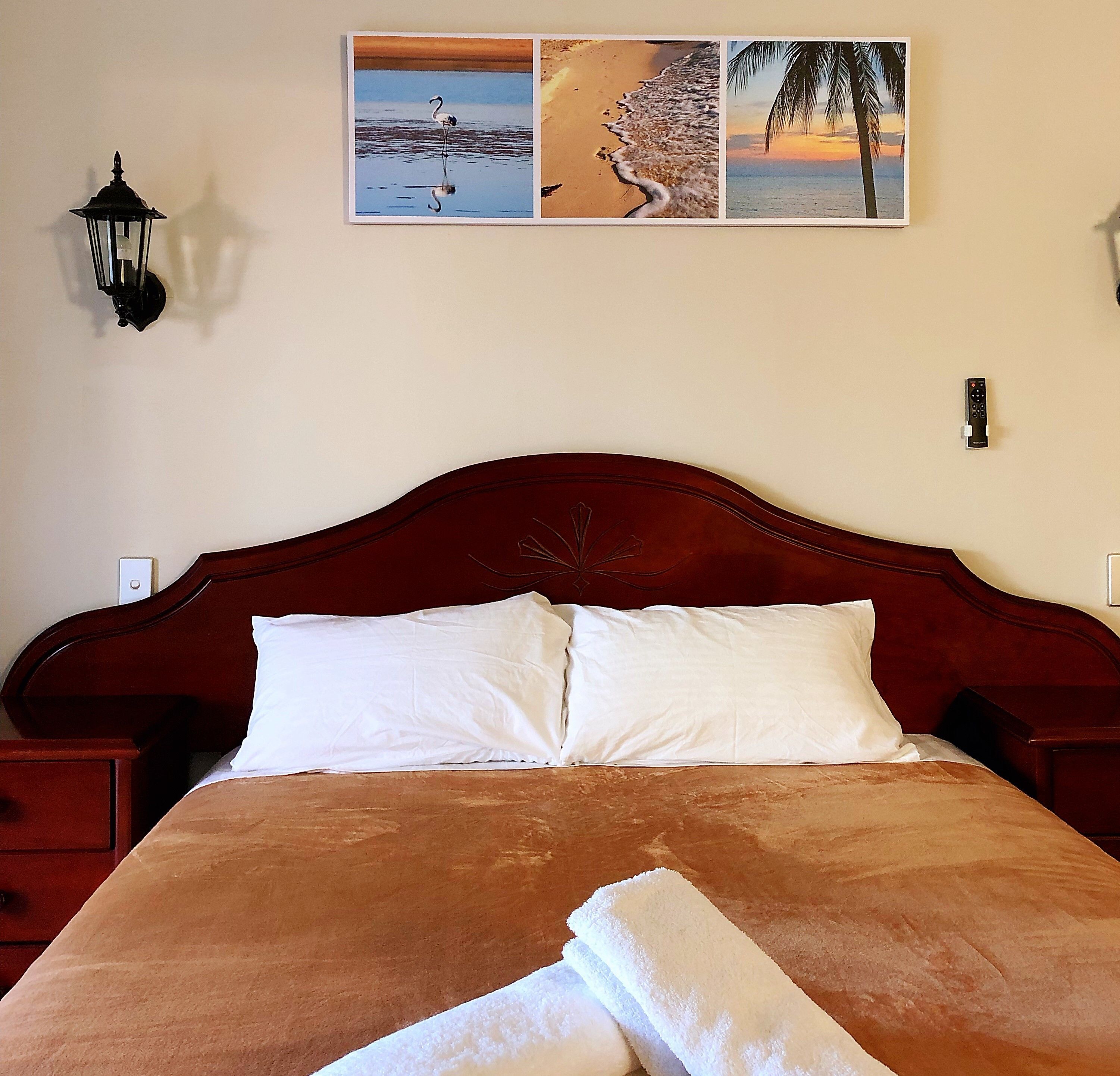 A bed with a wooden headboard, white pillows, and a brown bedspread. There are two wall-mounted light fixtures and a framed picture above the bed.