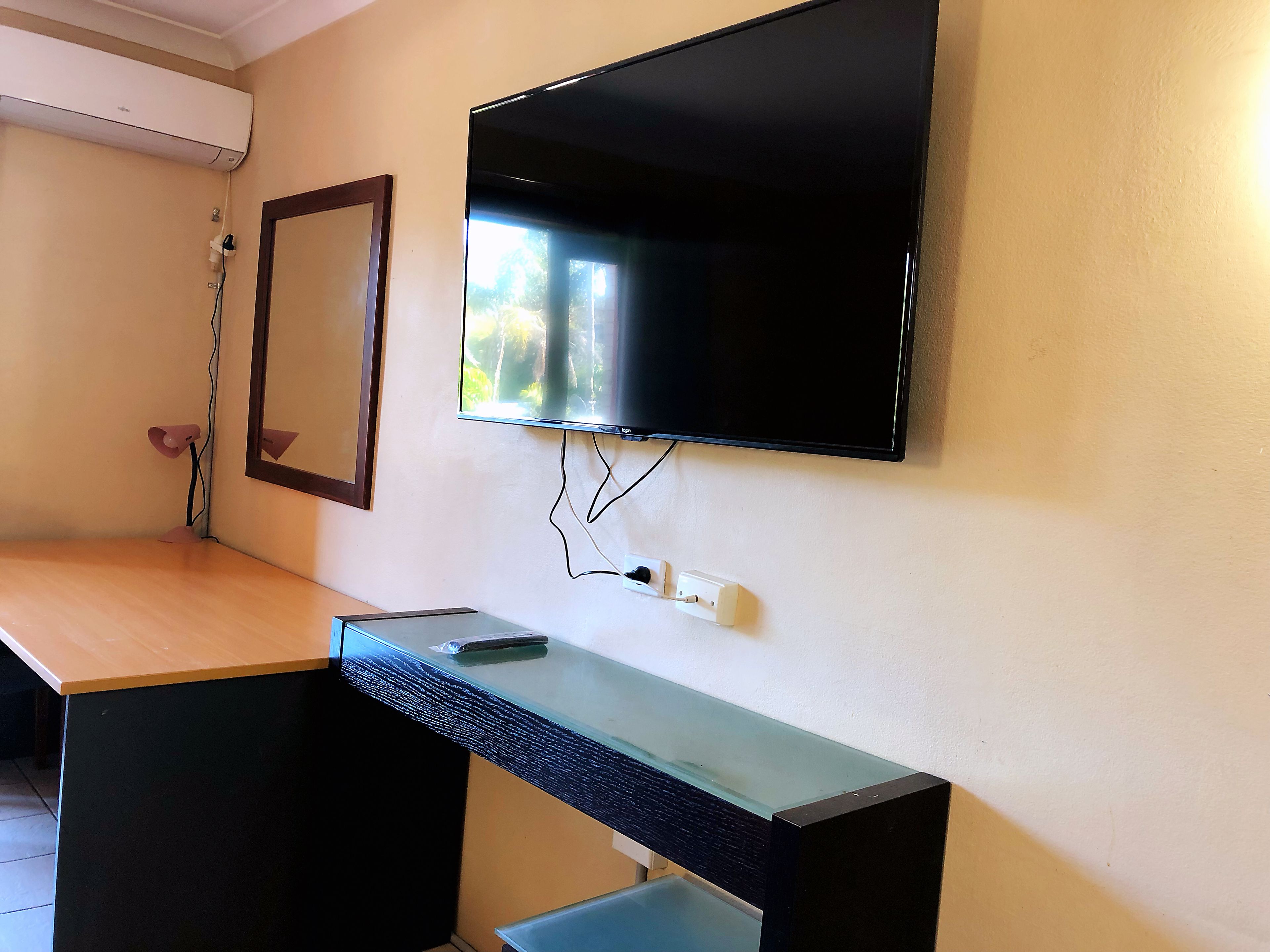 A room with a flat-screen TV mounted on the wall, a wooden desk with a glass top, and a mirror above the desk.
