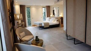 Une chambre d’hôtel moderne comprenant un lit, un coin salon avec un fauteuil et une petite table, une grande fenêtre et une porte coulissante donnant sur un balcon.