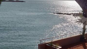 Um terraço com mesas ao ar livre, vista para o mar e uma árvore que oferece sombra.
