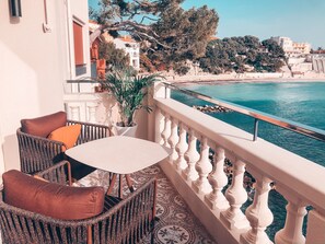 A balcony with wicker furniture, a potted plant, and a view of the sea and buildings.