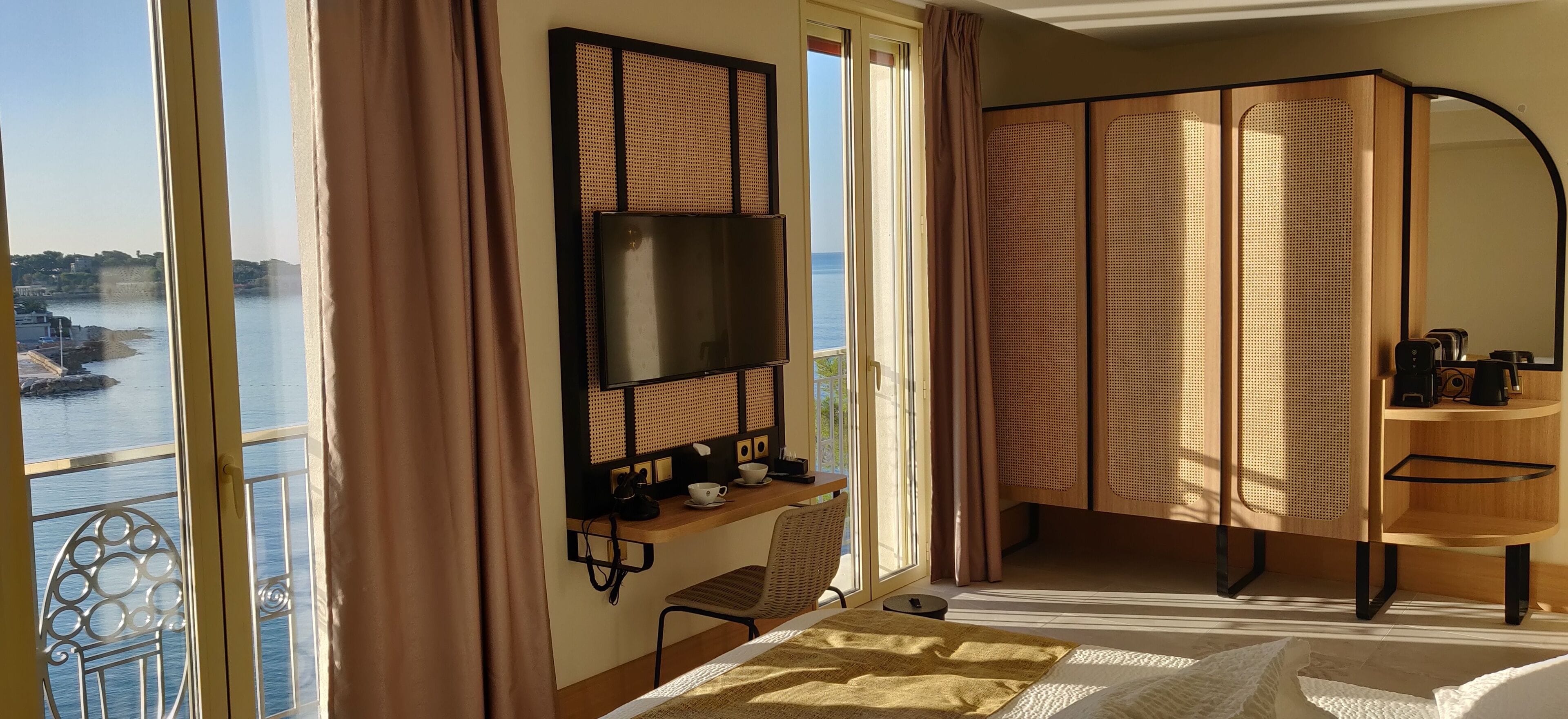 A modern hotel room with a large TV, a balcony with a view of the water, and a wooden cabinet with a kettle.