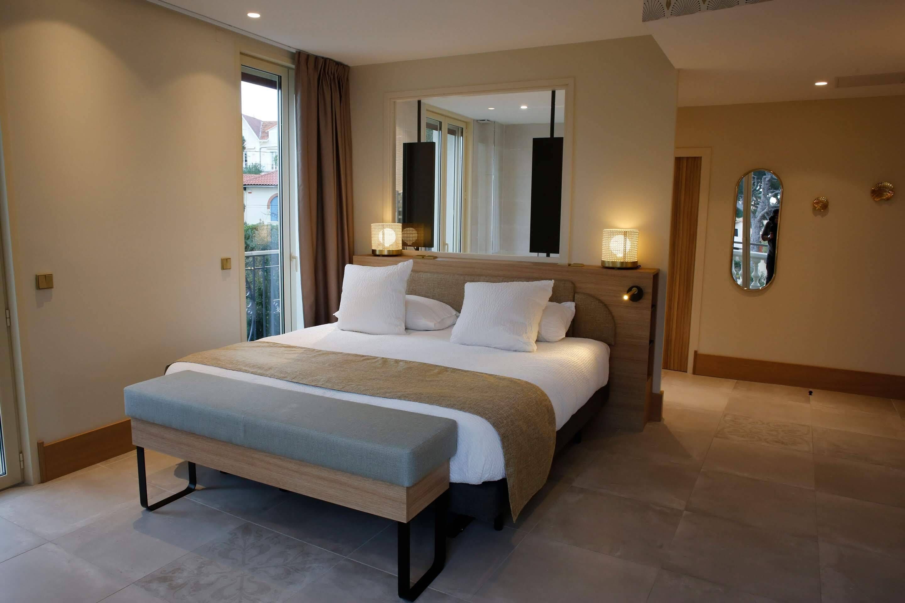 A modern hotel bedroom with a large bed, bedside lamps, a bench, and a large mirror.