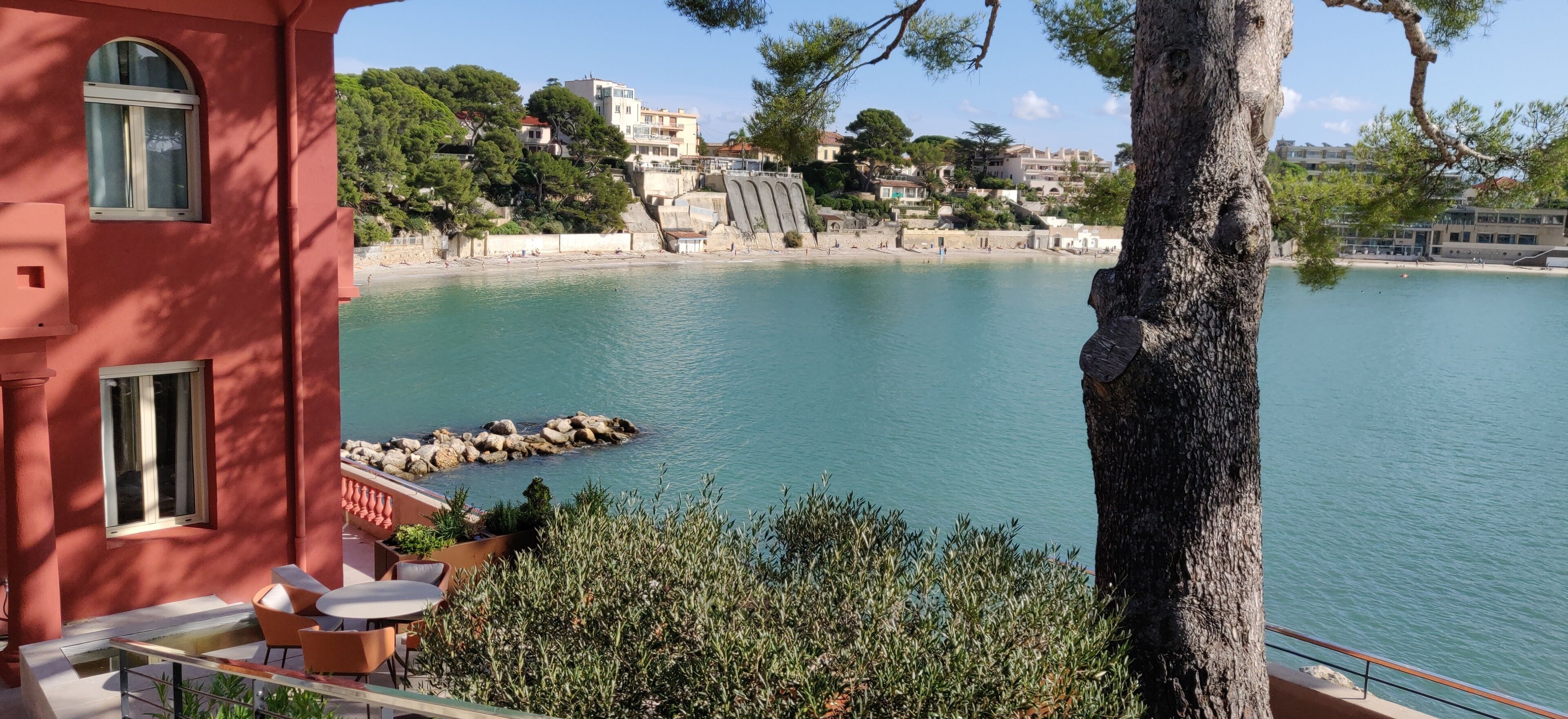 A coastal view with a red building, a rocky shoreline, and a clear blue sea.