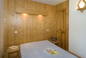 A small, cozy room with wooden walls and a single bed. There is a small table with a magazine and a suitcase on it. - Village de Gites le Colombier (Mende)