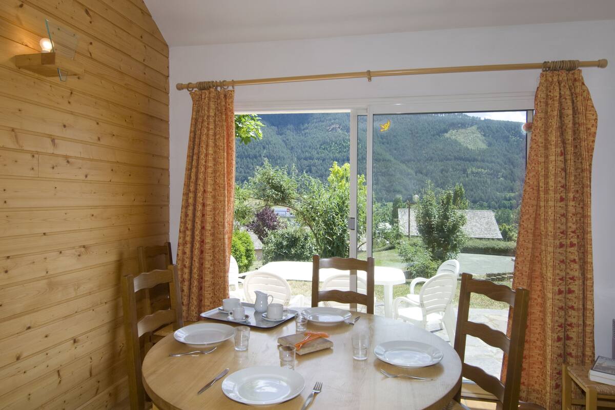 Un espace repas avec un décor intérieur en bois, une table dressée pour quatre personnes et une vue sur un paysage montagneux, visible à travers de grandes portes vitrées.
