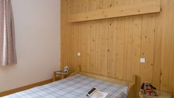 A room with wooden panel walls, a bed with a plaid bedspread, a small table with a teddy bear, and a wooden bench.