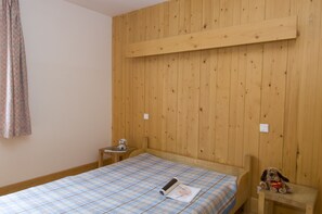 A room with wooden panel walls, a bed with a plaid bedspread, a small table with a teddy bear, and a wooden bench. - Village de Gites le Colombier (Mende)