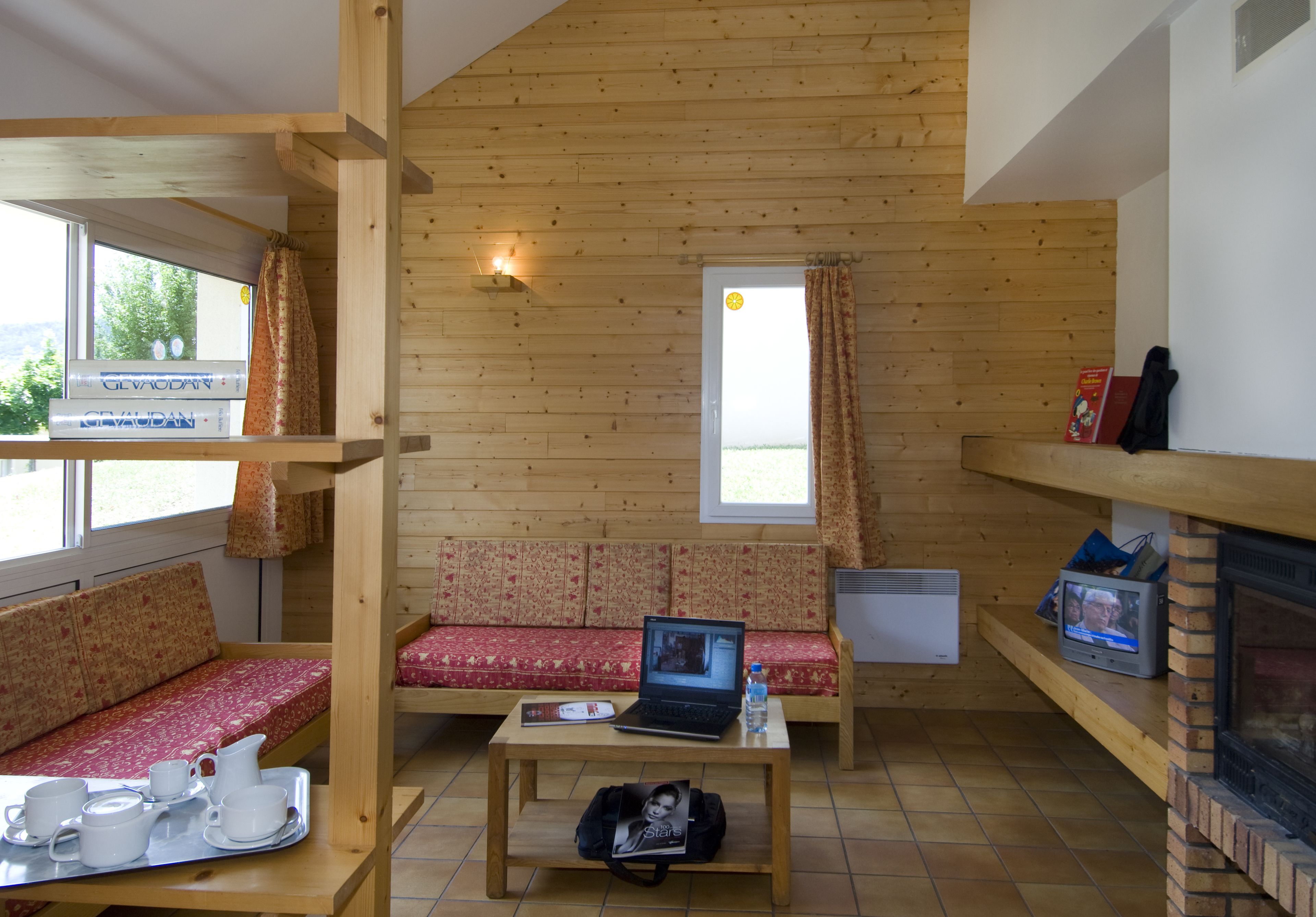 Un interior de cabaña acogedor, con paredes de madera, un sofá con cojines estampados, una mesa con una computadora portátil y una bandeja de té, y una chimenea.