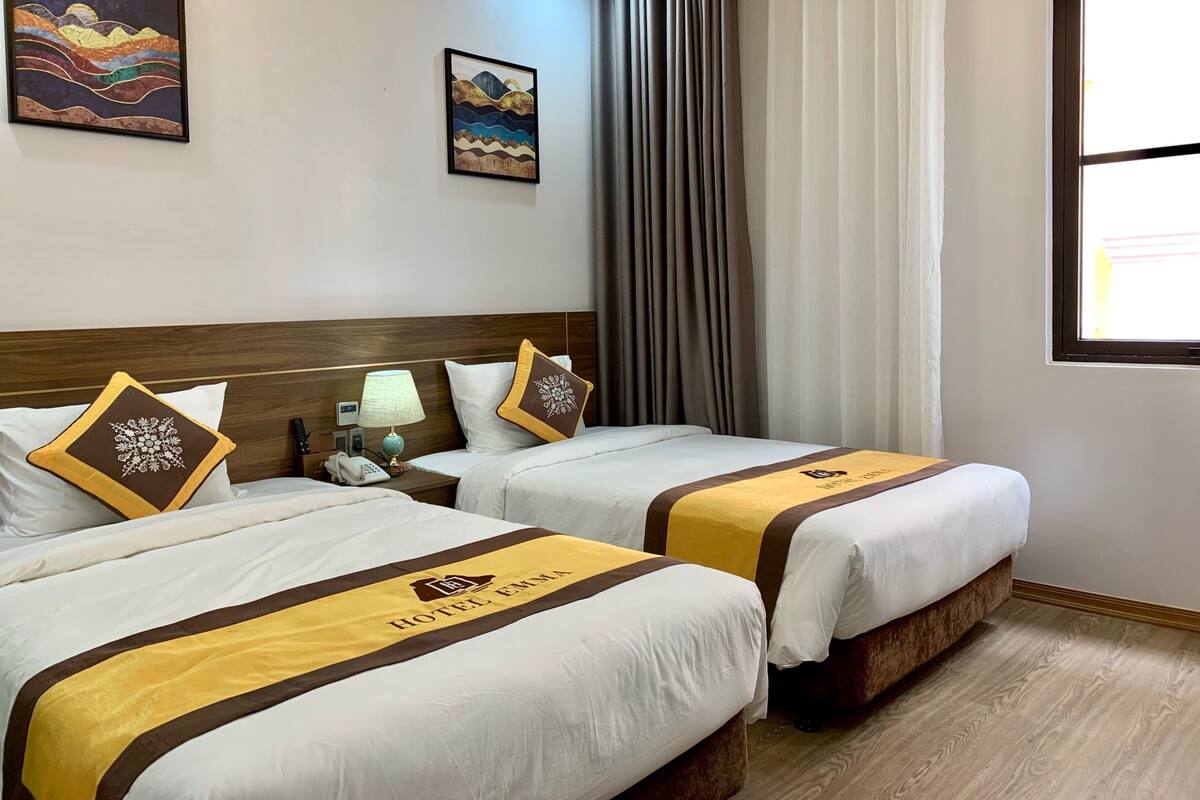 A hotel room with two beds, a wooden headboard, a nightstand with a lamp, a telephone, and two framed artworks on the wall.