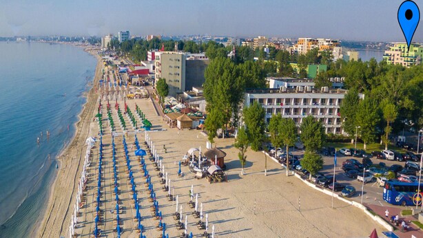 An aerial view of a beach with rows of umbrellas and beach chairs, a parking area, and buildings in the background. - Apart holiday Athena Mamaia (Constanta)