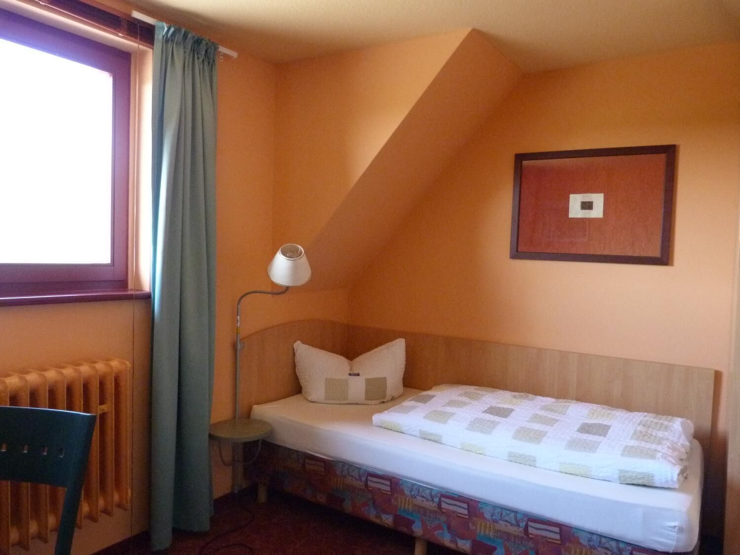 Ein kleines Zimmer mit einem Einzelbett, einem Holz-Kopfteil, einer Nachttischlampe, einem Heizkörper und einem Fenster mit Vorhängen.