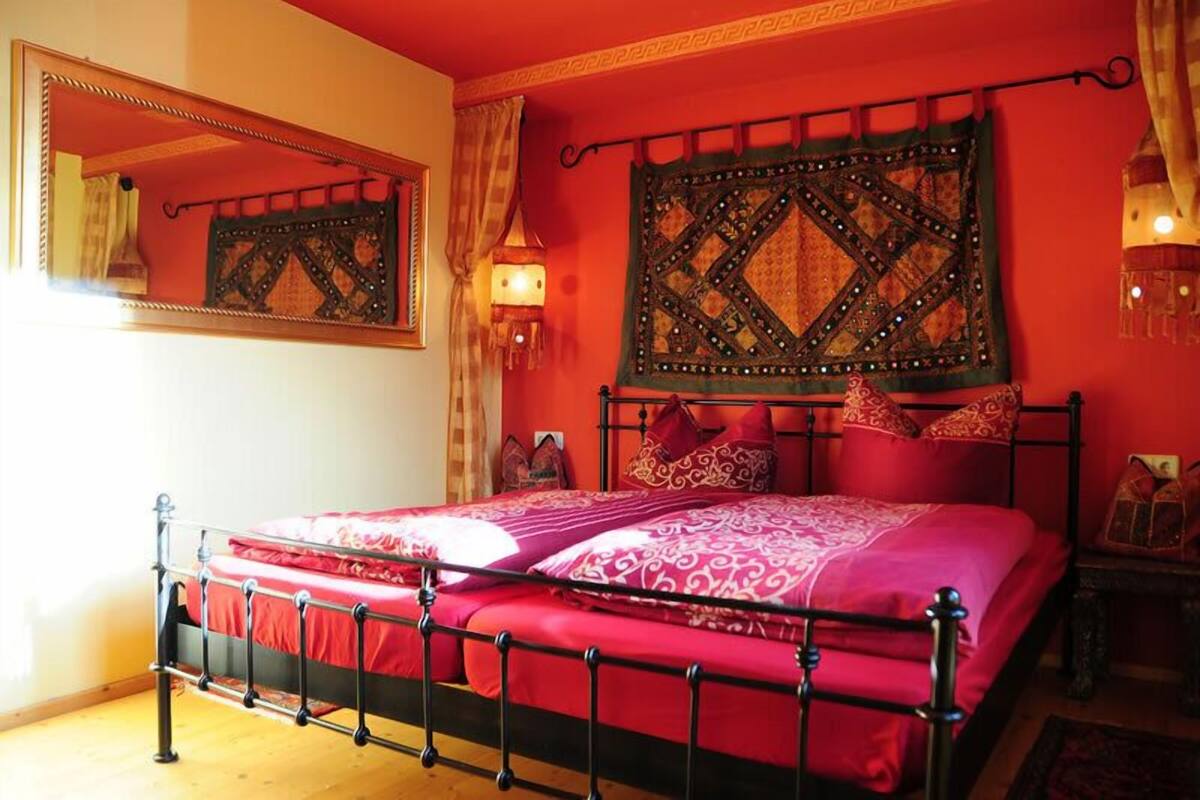 Ein Bett mit Metallgestell, roter Bettwäsche und einer dekorativen Wandweberei darüber.