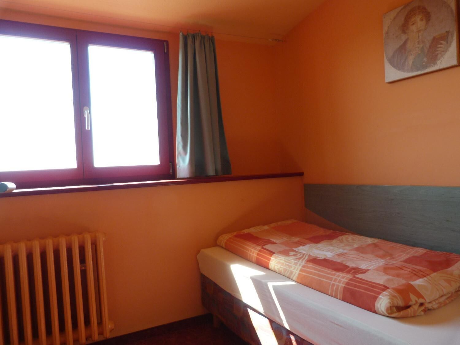 A small room with a single bed, a radiator, a window with curtains, and a painting on the wall.