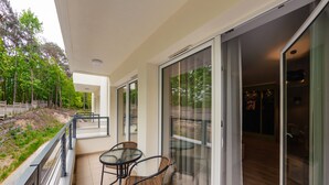 A balcony with a table and chairs, a glass door leading to an interior space, and a view of trees and a fence.