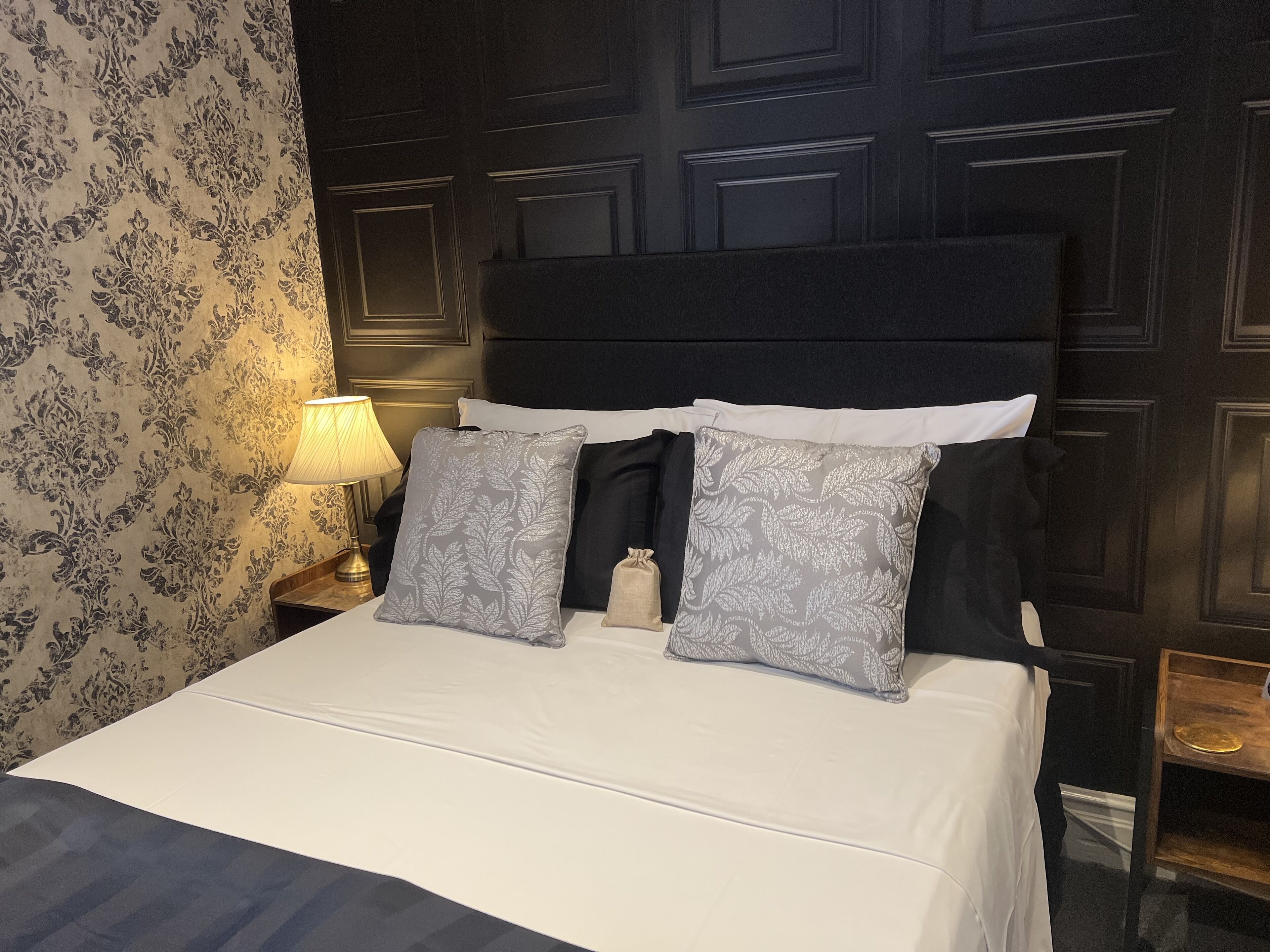 Un lit bien fait, avec du linge de lit blanc, deux coussins gris à motifs et une tête de lit noire.
