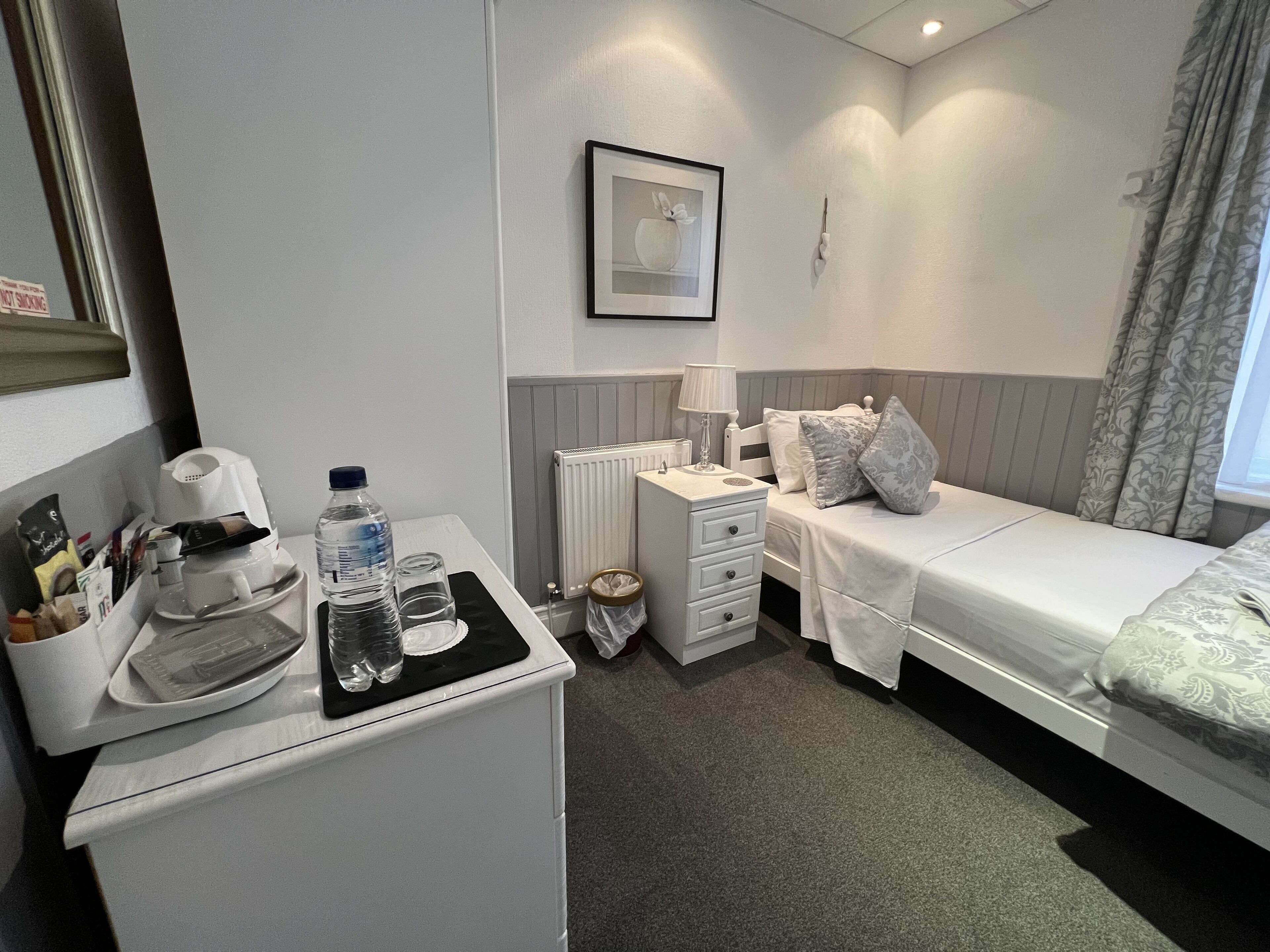Habitación de hotel con cama, mesita de noche, lámpara y una pequeña zona de kitchenette con cafetera y botella de agua.