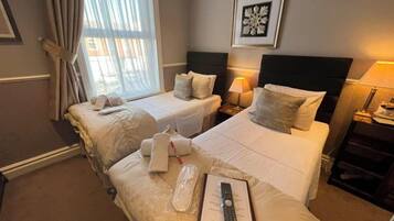 A hotel room with two beds, a window with curtains, a framed artwork on the wall, and a bedside table with a lamp.