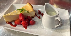 A dessert plate with a slice of cheesecake, raspberries, and a small pitcher of sauce.