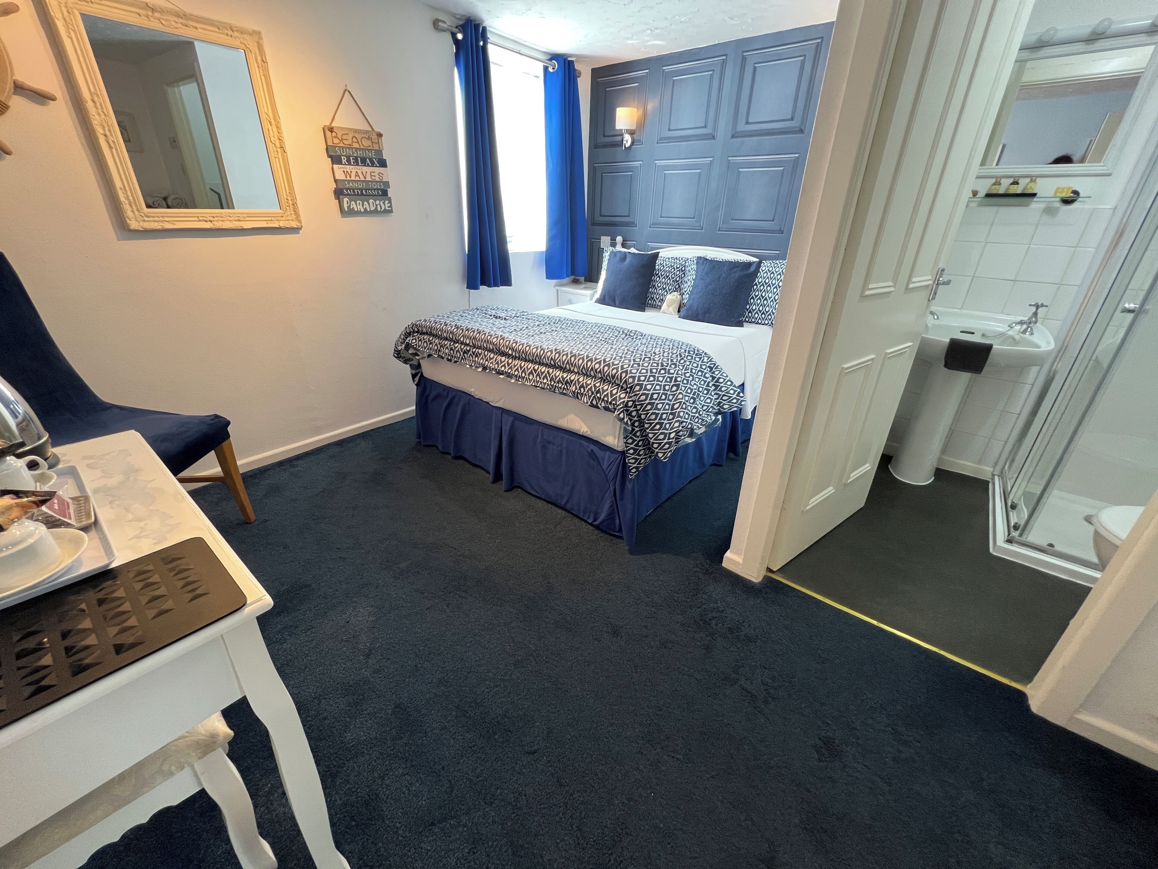 A bedroom with a bed, a chair, a table, a mirror, and a bathroom with a sink and shower.