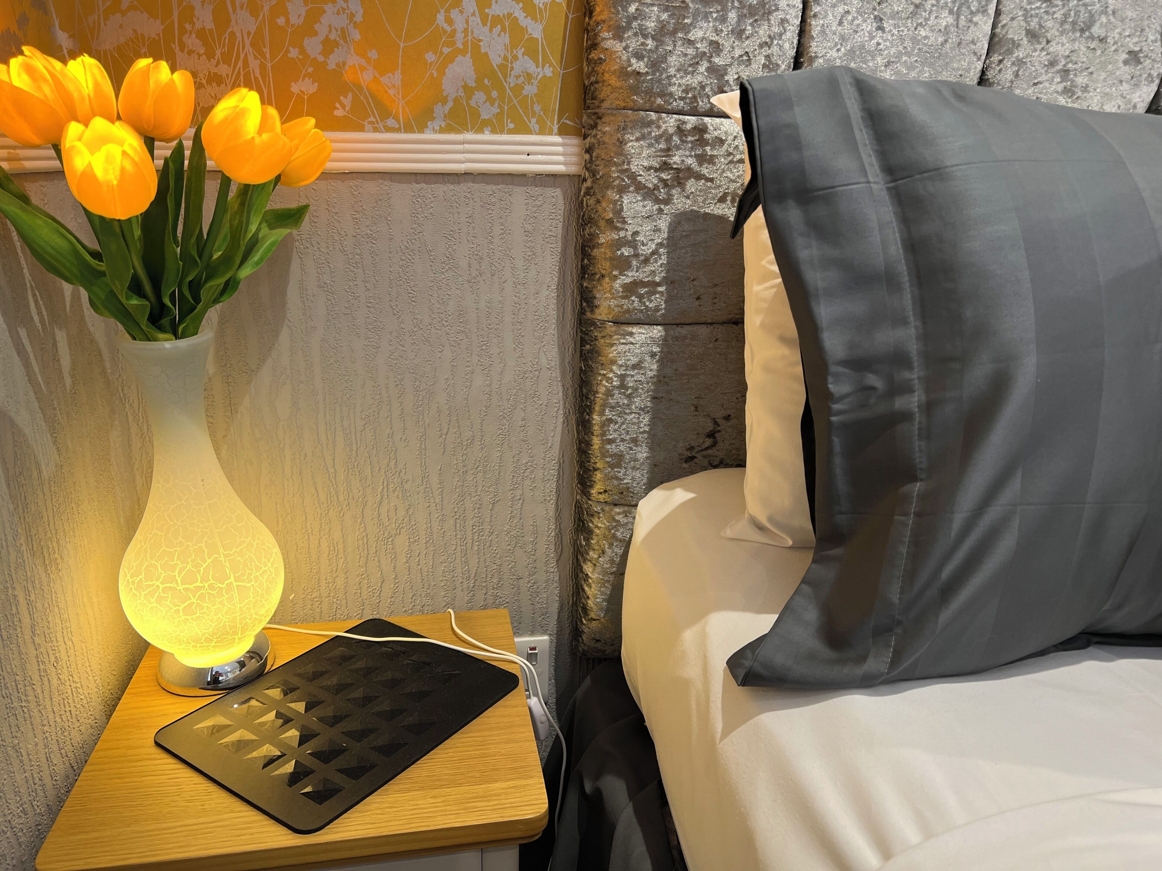 Una mesita de noche con una lámpara, un adaptador de corriente y un jarrón decorativo con flores.