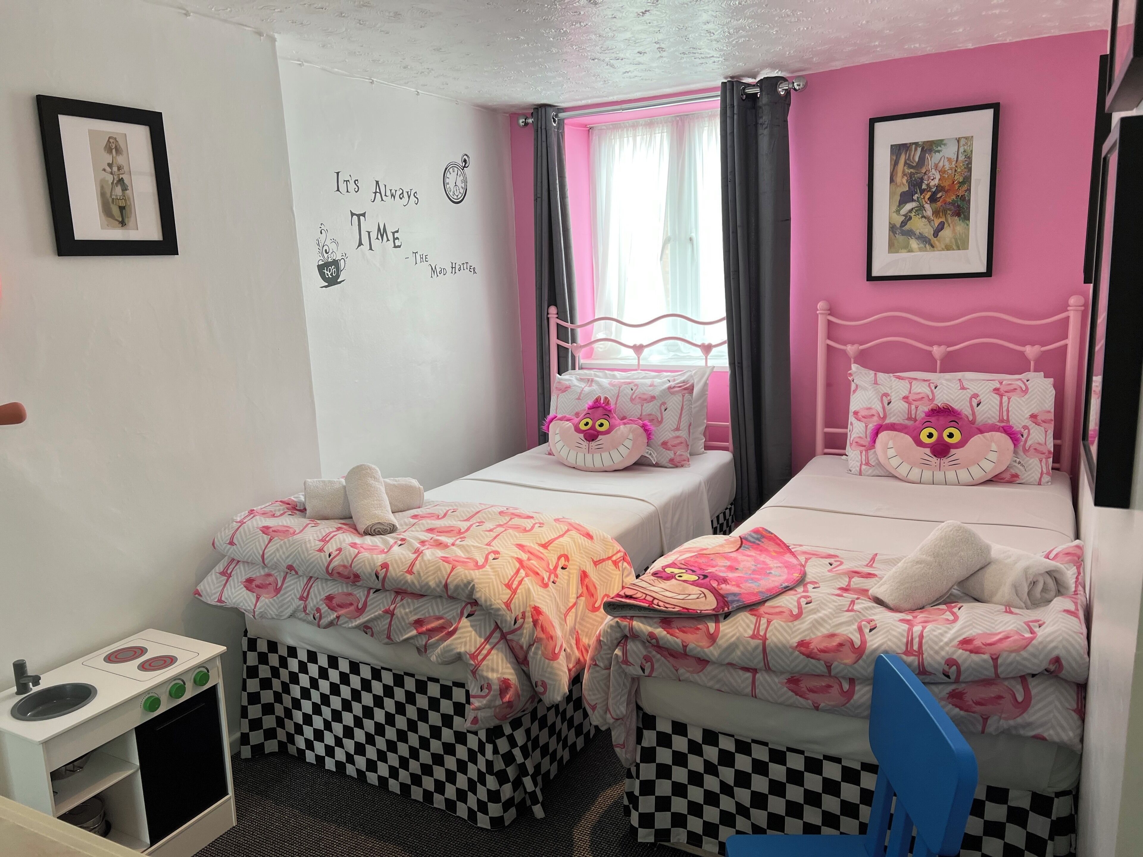Un dormitorio con dos camas, cada una con ropa de cama temática de flamencos rosas, una kitchenette pequeña y una pared con una cita y un reloj.