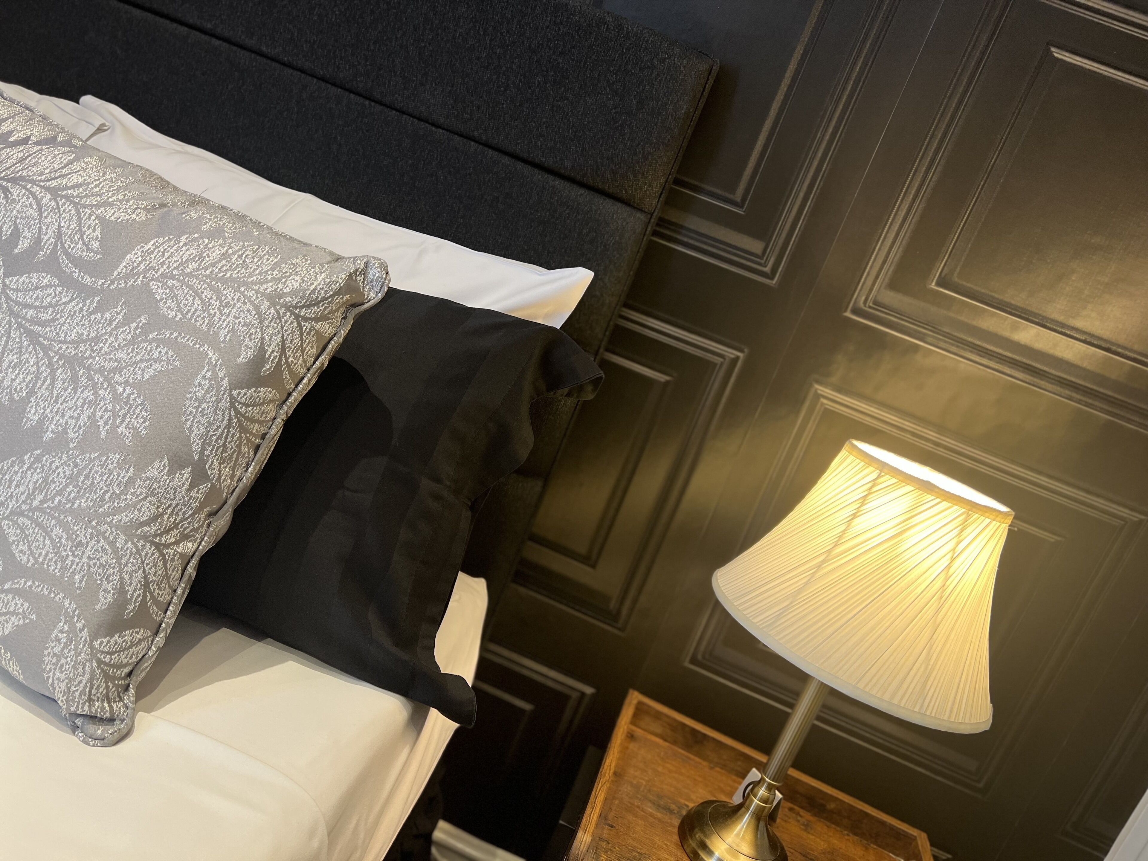 A neatly made bed with a patterned pillow and a black pillow, a bedside lamp, and dark wall paneling.
