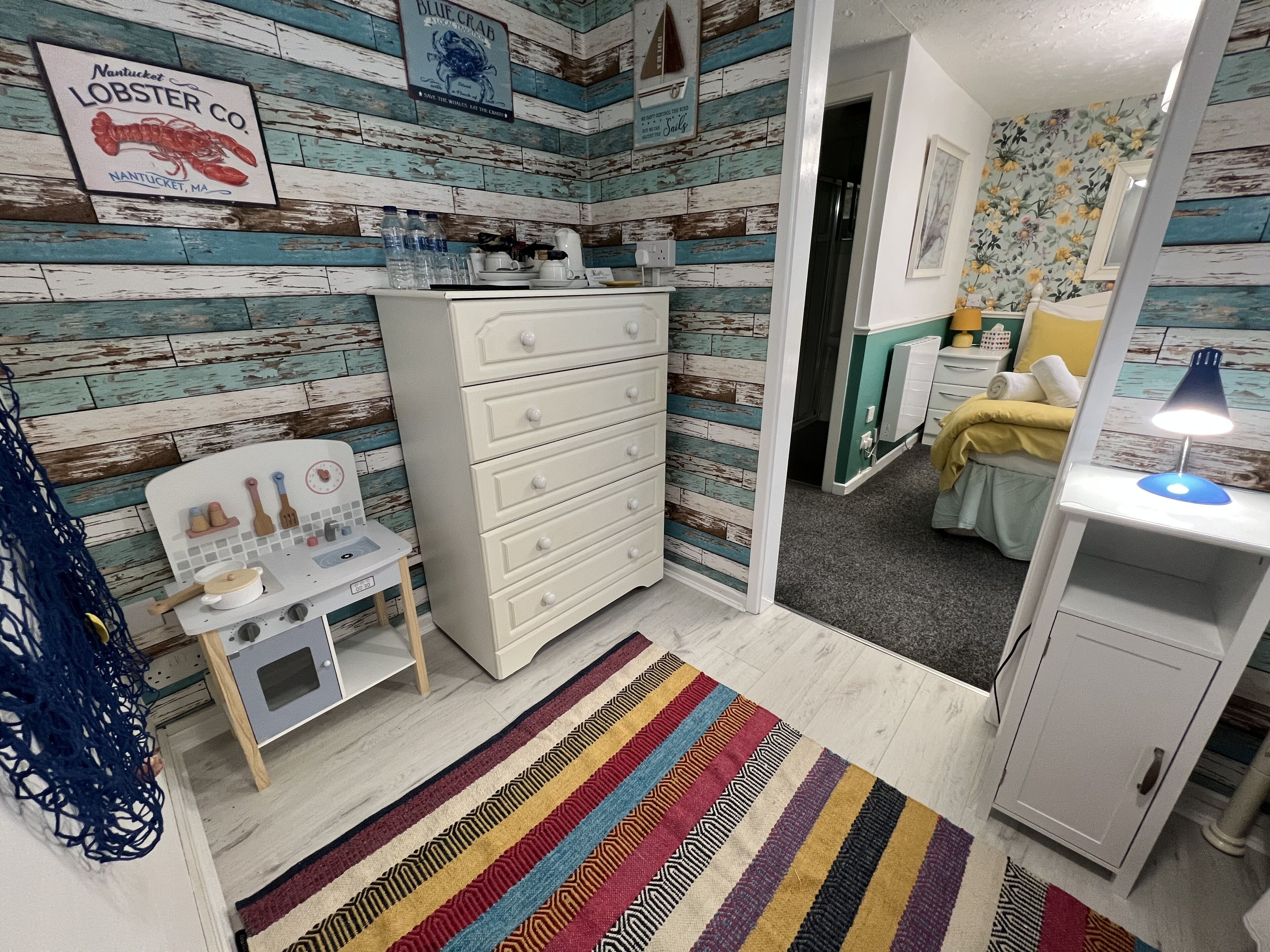 A room with a wooden plank wall, a white dresser, a small desk with kitchen utensils, a colorful striped rug, and a bed with a yellow blanket.