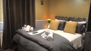 Una cama bien hecha con un edredón oscuro, almohadas y una toalla enrollada. En la mesita de noche hay un pequeño florero con flores amarillas.