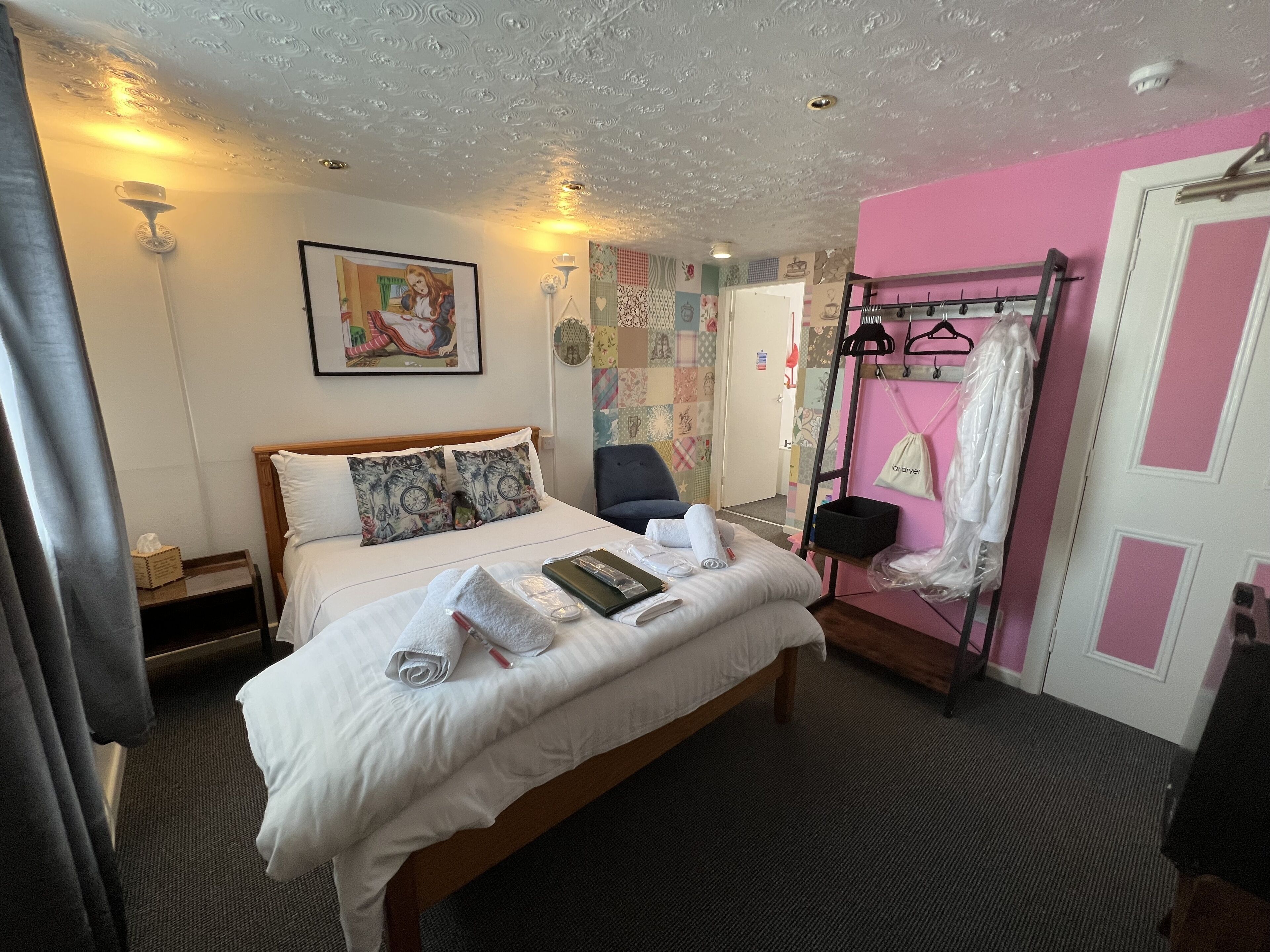 Een hotelkamer met een netjes opgemaakt bed, een stoel, een garderobe met daarin kleding en een muur met een kleurrijk patroon.