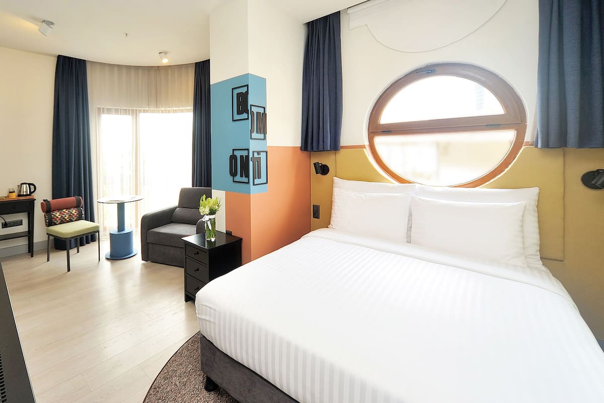 A hotel room with a large bed, a desk with a chair, a small table with a vase, and a window with curtains.