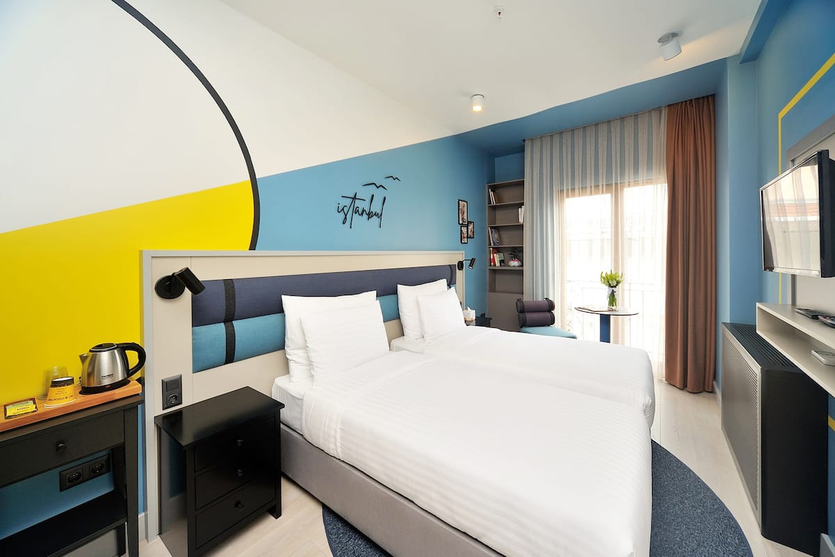 A hotel room with a bed, bedside tables, a TV, and a wall with a city name and a bird.