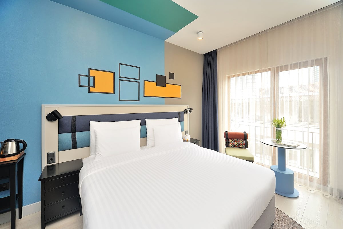 A modern hotel room with a large bed, a nightstand, a small table with a plant, and a view of the city through large windows.