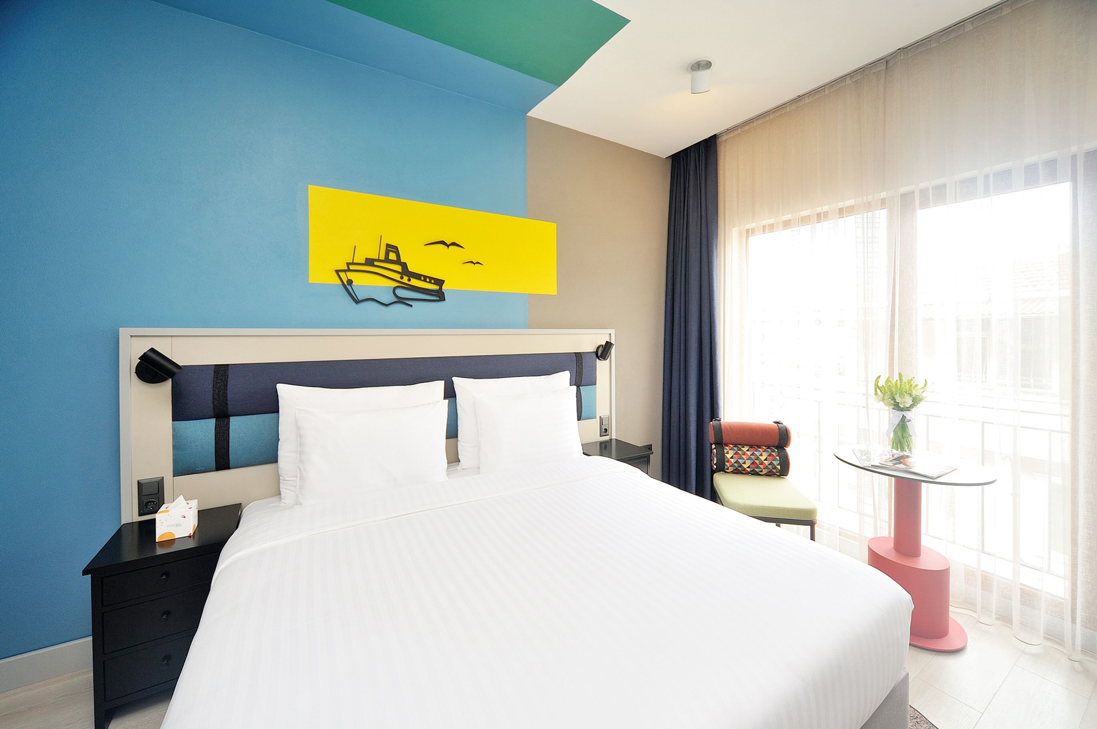 A hotel room with a large bed, a nightstand, a chair, a small table with a plant, and a window with curtains.