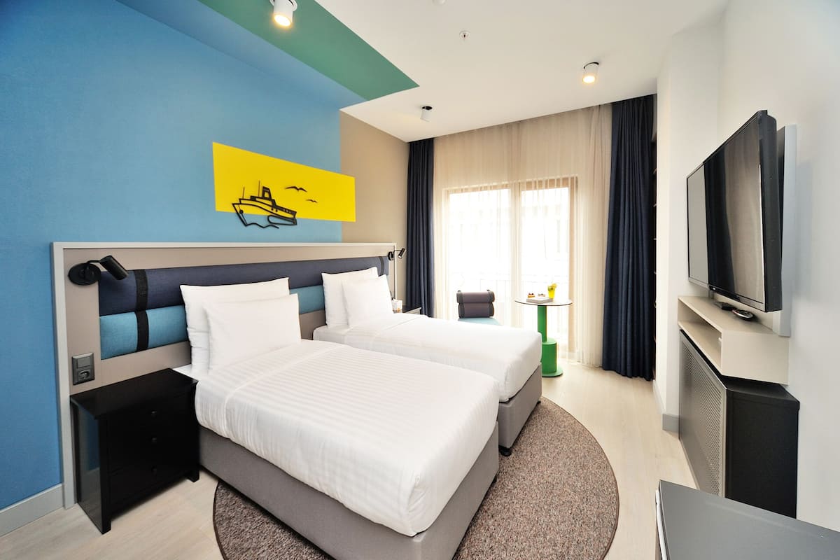 A hotel room with two beds, a TV, a painting, and a window with curtains.