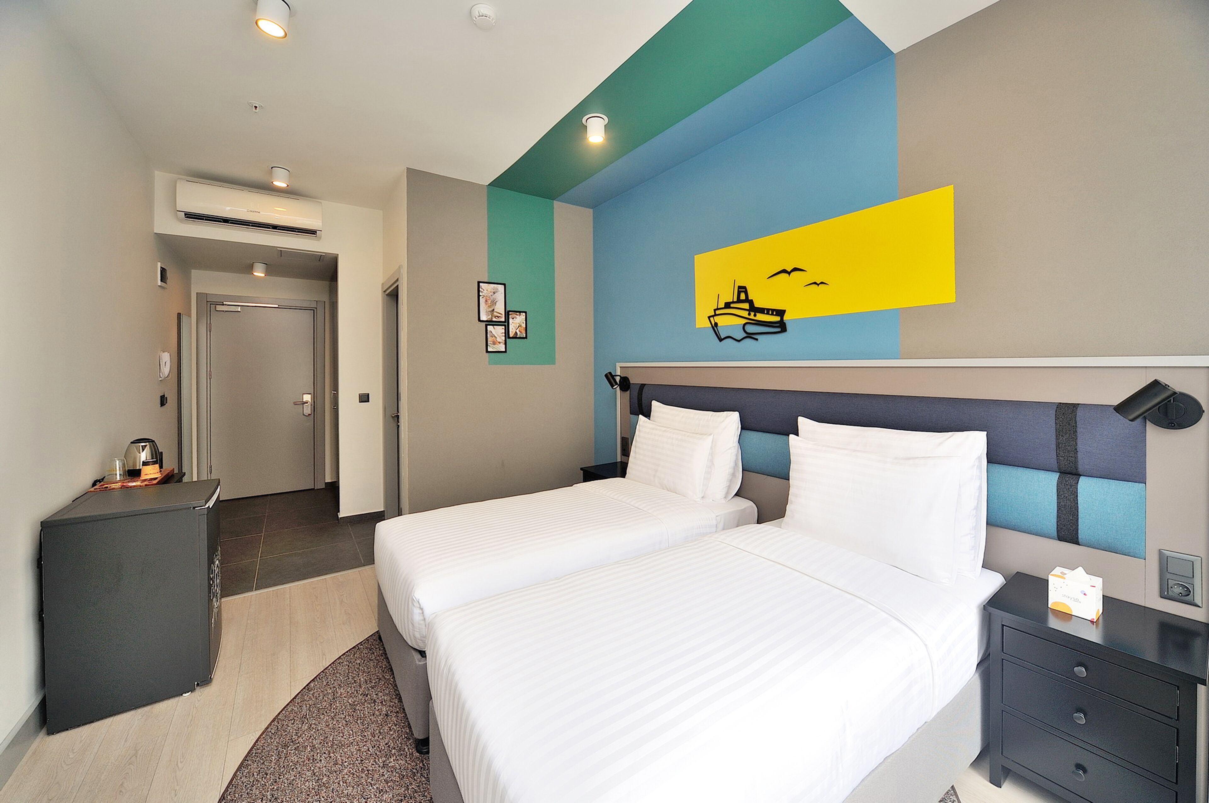 A hotel room with two beds, a small desk, and a wall-mounted air conditioner.