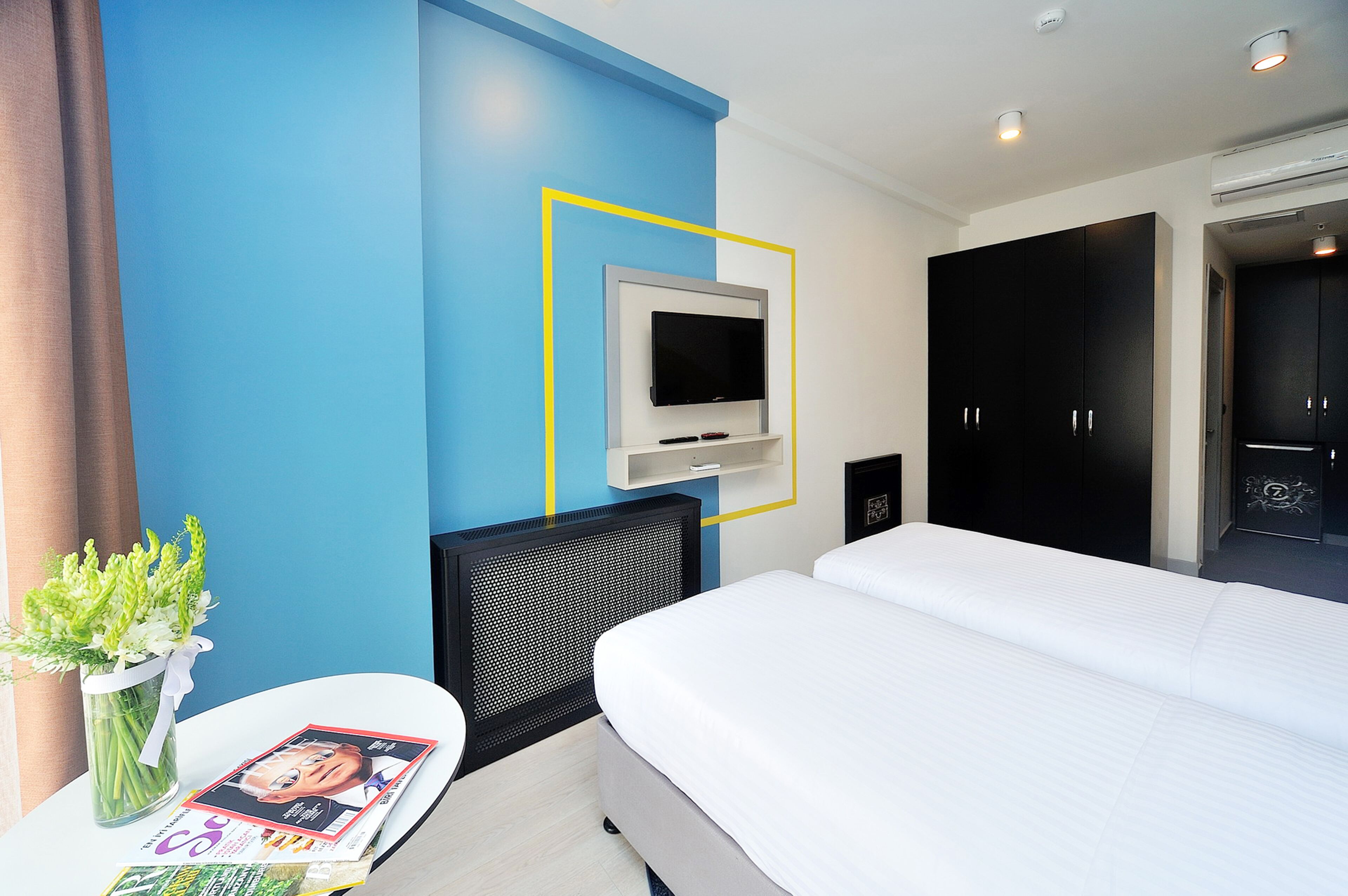 A hotel room with a bed, a TV mounted on the wall, a desk with a magazine, and a vase with flowers.