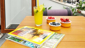 Una mesa de madera con una revista, un vaso de bebida amarilla con una rodaja de limón, y cuencos de bayas en un balcón con una valla de metal y edificios al fondo.