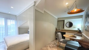 Una habitación de hotel moderna con cama, zona de comedor con mesa y sillas, mostrador de cocina y un amplio ventanal con cortinas.
